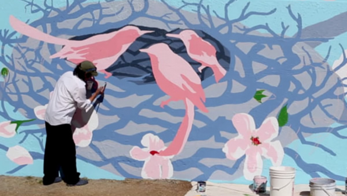 A person painting an outdoor mural depicting a nest with birds and flowers