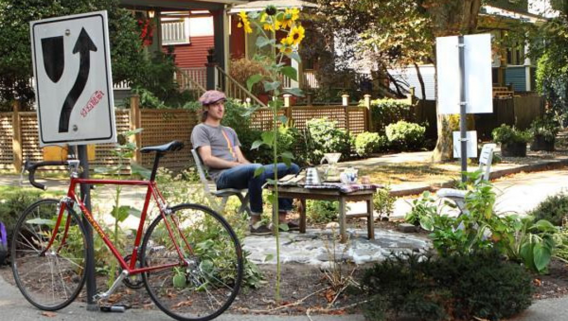 Person sitting in the middle of a roundabout next to a table with refreshments. Flowers and a bicycle are in the foreground.