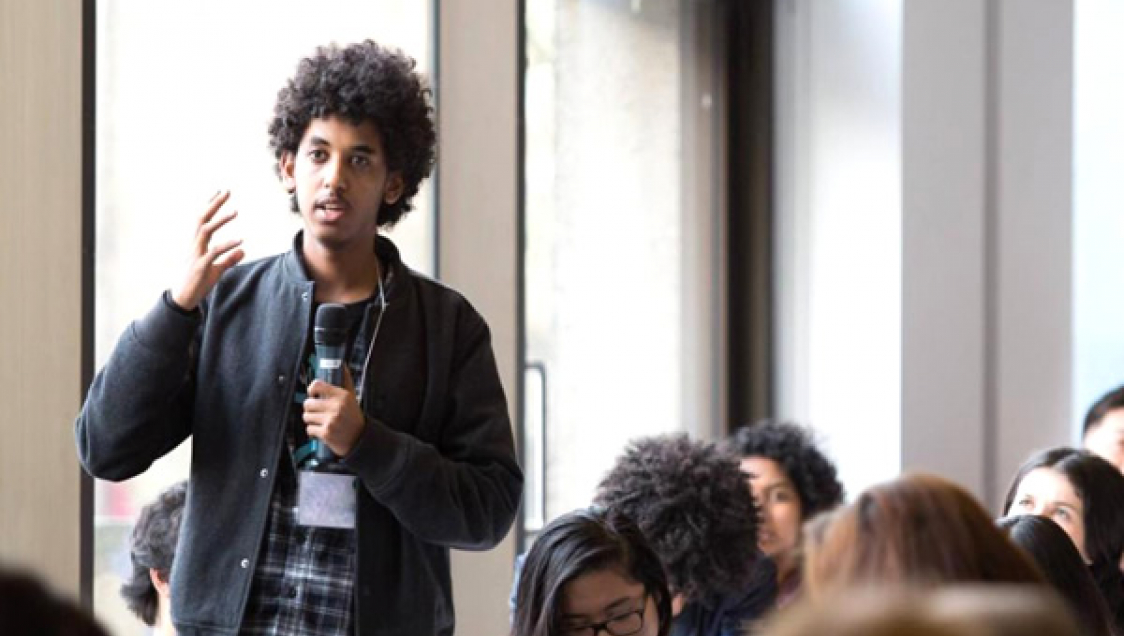 A young person speaking into a microphone, standing, with others seated nearby.