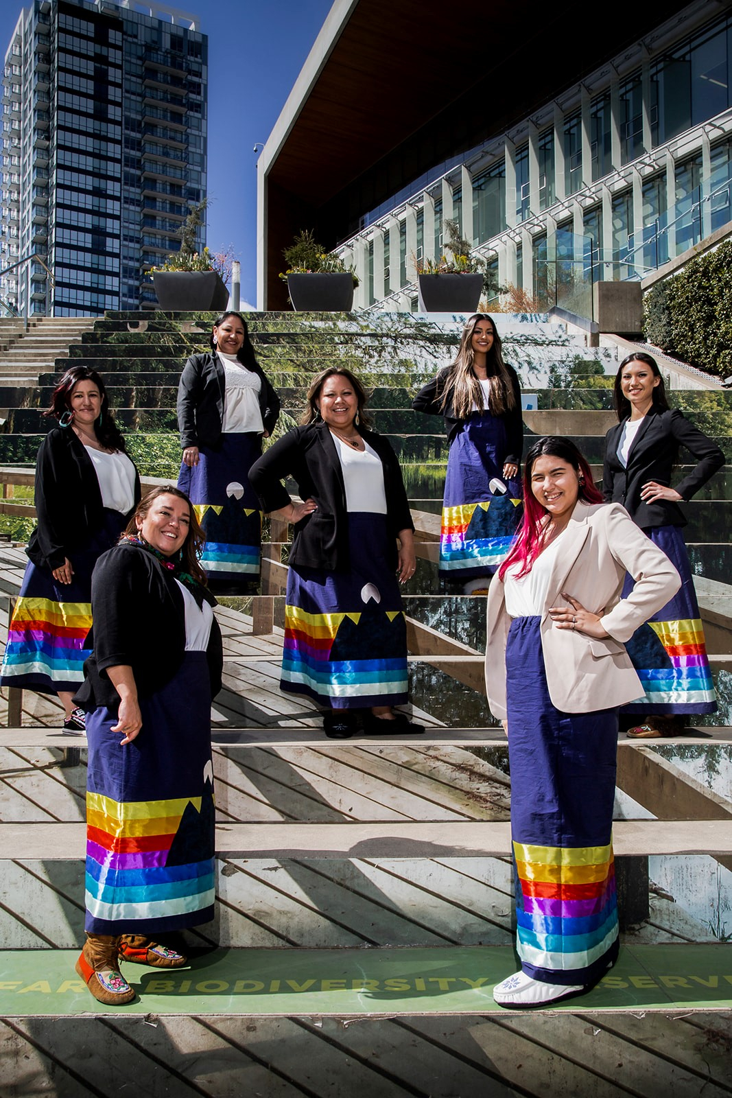 Several Indigenous women smiling and wearing ribbon skirts stand spaced apart on steps.
