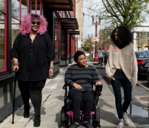Three women walking down the street smiling. One woman has a cane and another is in a wheelchair.
