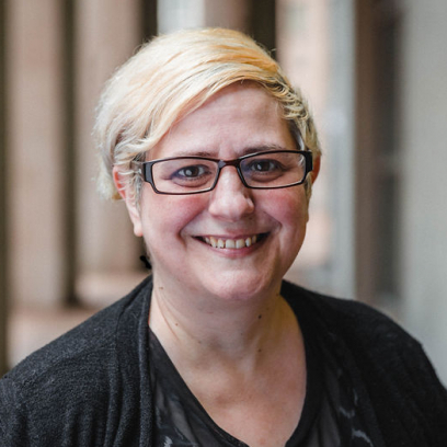 Headshot of Twila Schembri. She has short light blonde hair and brown eyes and is wearing square framed glasses and a dark top and cardigan.