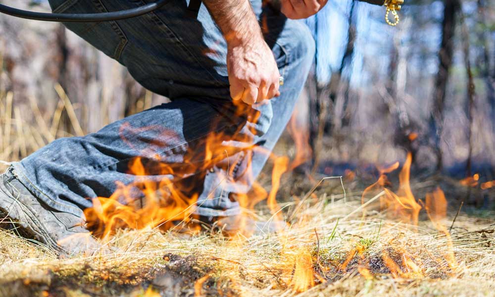Waist down shot of a man in jeans standing near dry brush that has caught fire