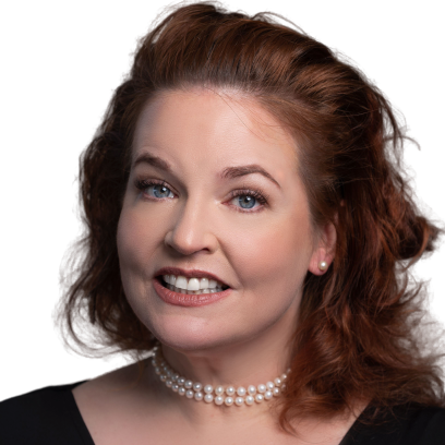 A smiling woman with light eyes and auburn hair wearing a pearl necklace and dark blouse
