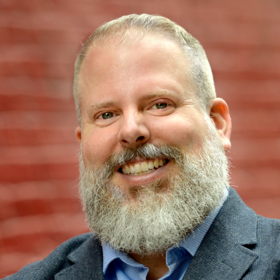 Headshot of Mike Conroy, a smiling man with light eyes, grey hair and a bushy beard wearing a blue shirt and grey suit jacket.