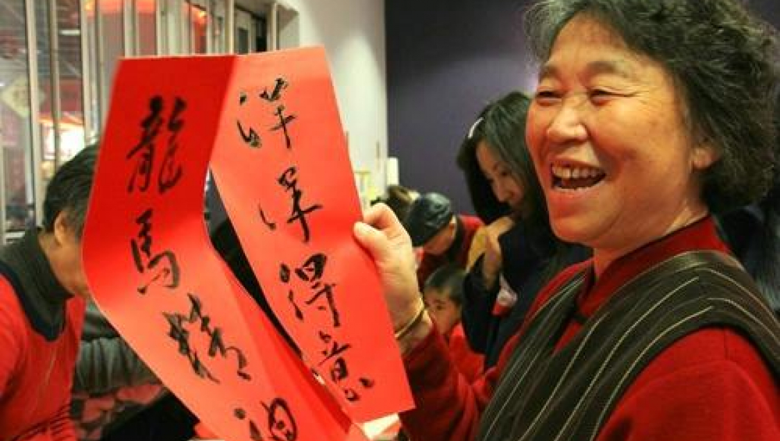 An older asian woman smiling and holding two pieces of red paper with chinese calligraphy on them in ink