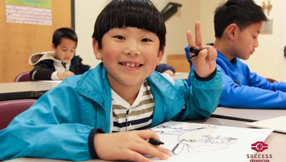 A little asian boy in a blue jacket and holding a sharpie smiles at the camera and makes a peace sign with his hand, logo in the lower right corner reads "Success Foundation"