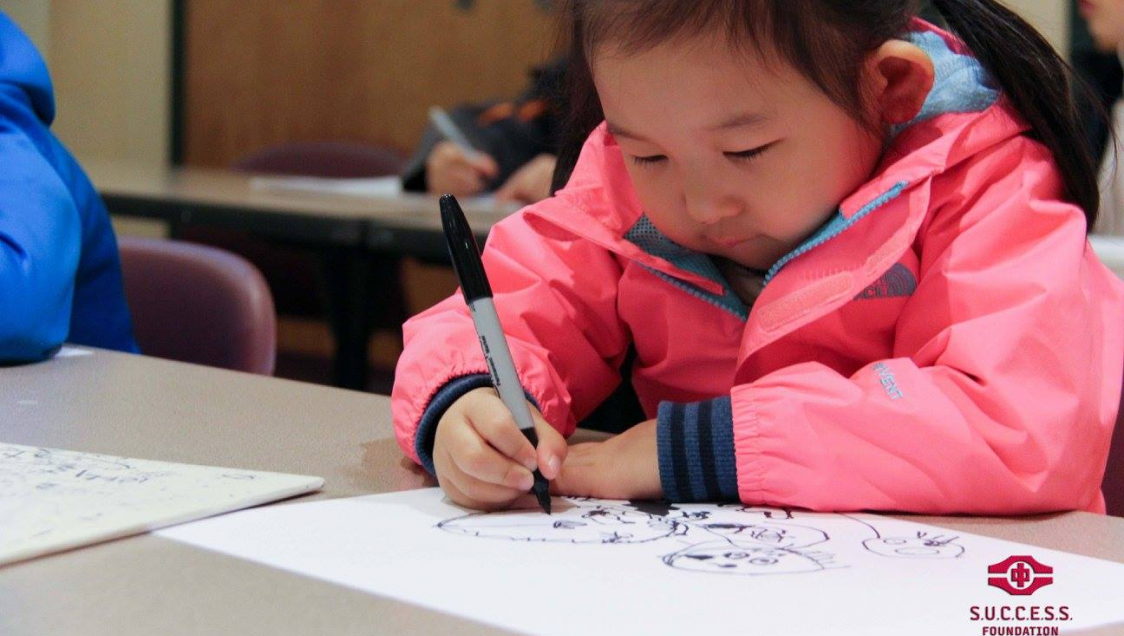 A little asian girl in a pink jacket drawing on paper with a sharpie, logo in the bottom right corner reads "Success Foundation"