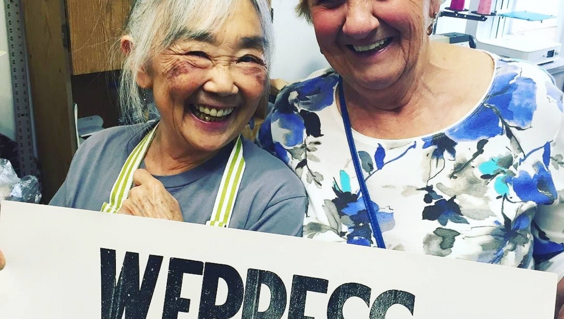 Two smiling elderly women holding a sign that reads "WePress"