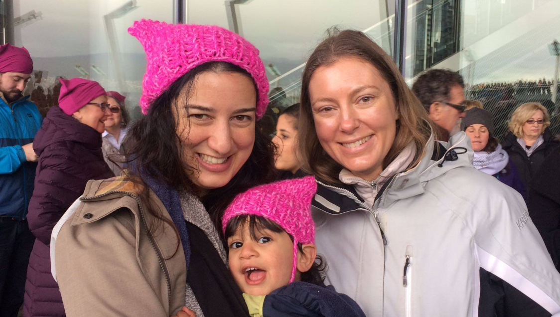 A dark haired woman holding a little girl and both wearing knitted pink hats stand next to another woman in a grey jacket and smile