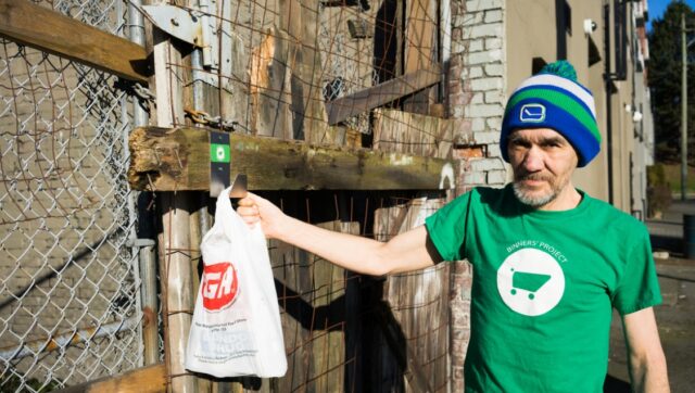 An elderly man in a toque and green shirt with a white shopping cart graphic hangs a plastic bag on hook attached to a wooden fence