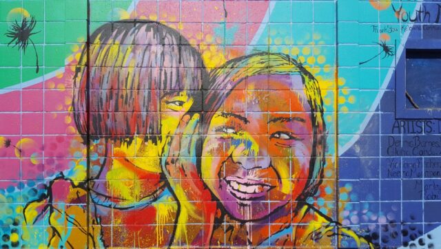 A colourful spraypainted mural of a child whispering to another smiling child