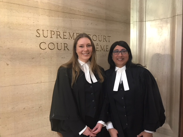 Two women in formal court attire of dark vests and cloaks, and white neckties, standing before a wall with the words "Supreme Court" carved into the stone