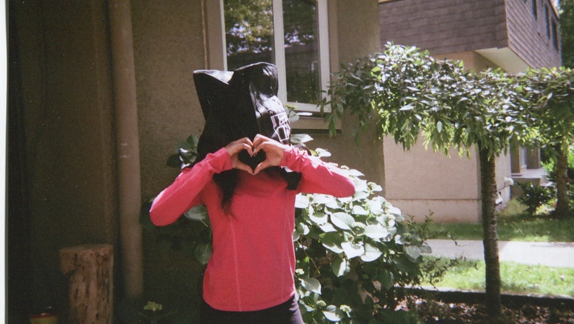 A woman in a pink long sleeved top with a black bag over her head stands outside of a house and makes a heart-shape with her hands