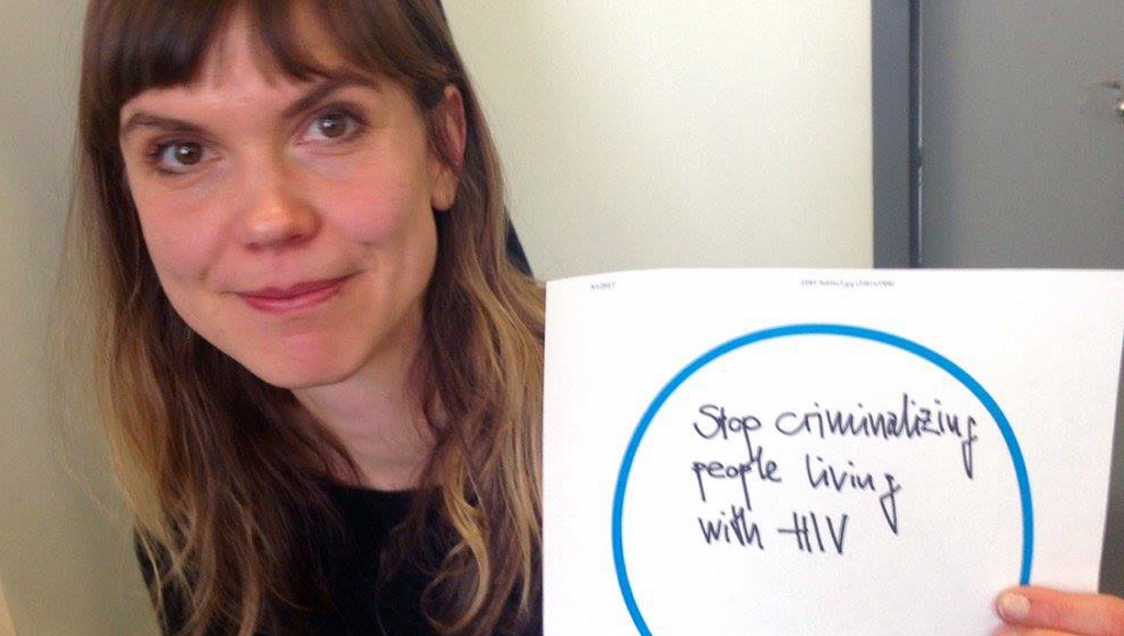 A smiling woman holding up a piece of paper with "Stop criminalizing people living with HIV" written on it