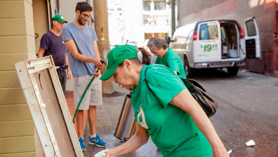 A diverse group of people in green shirts with a white shopping cart graphic washing various things in the street