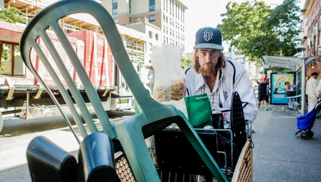 A white man with long brown hair wearing a basseball cap stands with a shopping cart full of plastic items and chairs