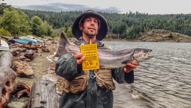 A man in a waterproof coat and bucket hat, holding up a fish and a yellow oil spill card on the coastline