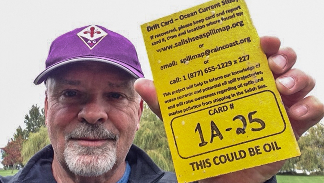 An older white man with a goatee and mustache wearing a purple ballcap, holding up an oil spill identification card 