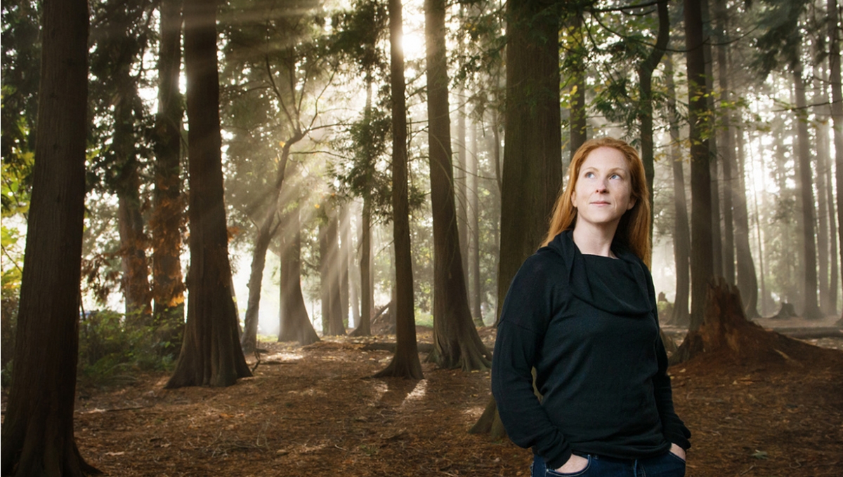 A white woman with red hair wearing a dark long sleeved top and jeans stands in a sunlight wooded forest