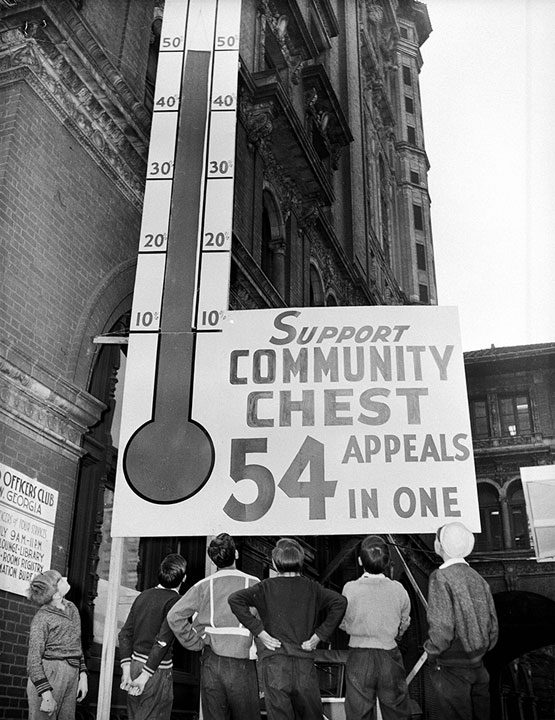 Large poster showing funds raised for a community chest appeal towers over a group of men and boys in a black and white photo.