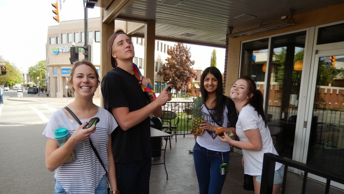 Four diverse youth striking poses in the patio area of a cafe, holding cupcakes and a pineapple