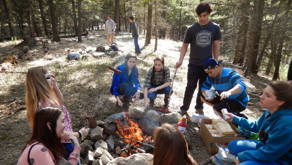 A diverse group of youth around a campfire in a wooded forest area, grilling hot dogs together over the flames