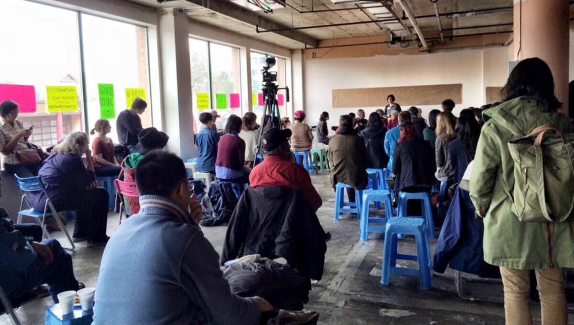 A group of people sitting in a bare and concrete building listening to a public speaker