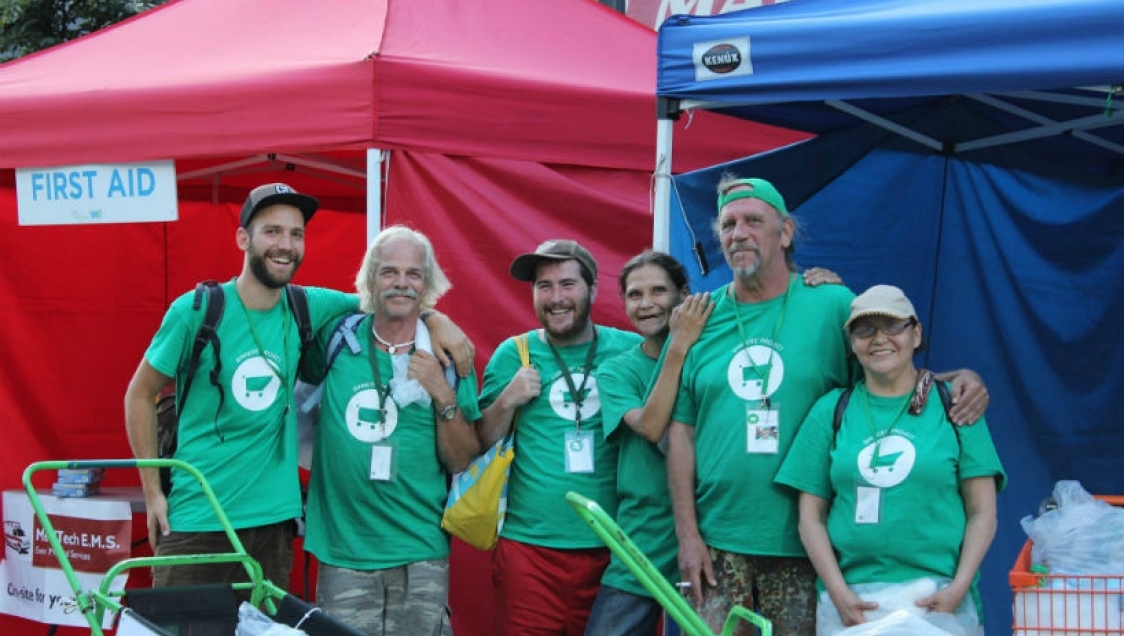 A diverse group of people in green shirts with a white shopping cart graphic standing in front of stalls with tents and smiling