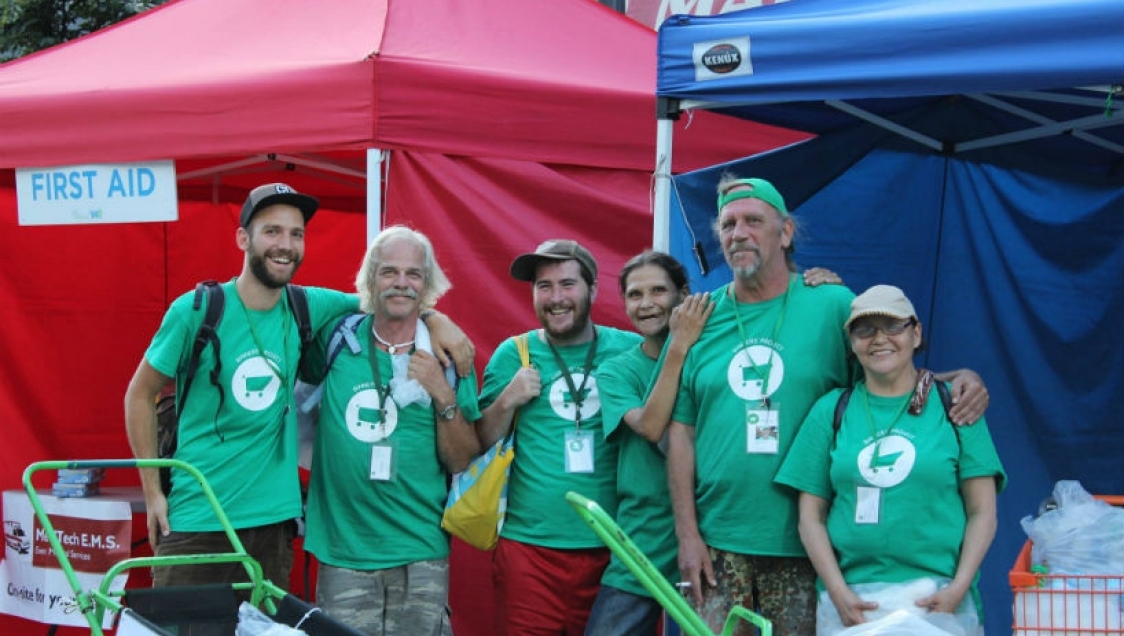 binners_project_4 A diverse group of people in green shirts with a white shopping cart graphic standing in front of stalls with tents and smiling