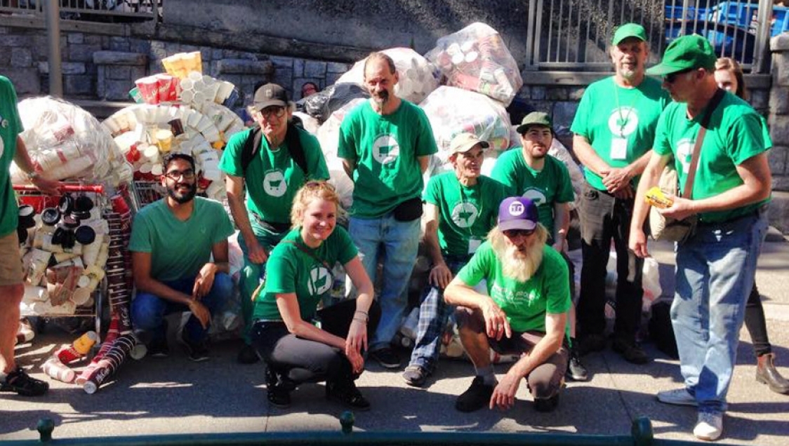 A diverse group of people in green shirts with a white shopping cart graphic pose with shopping carts full of bags of paper cups behind them