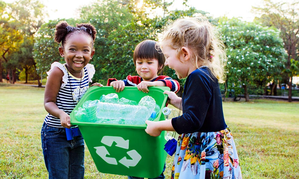 Three young children of diverse backgrounds holding a green recycling bin full of empty plastic bottles and smiling