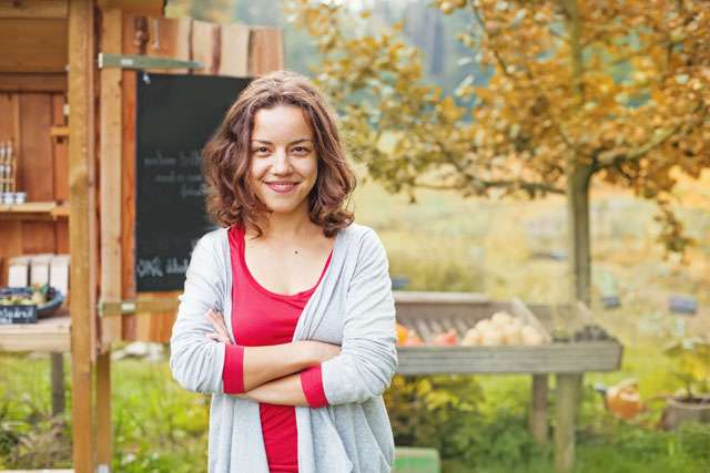 Smiling woman with shoulder length brown hair standing in front of a wooden vegetable stall and chalkboard, arms crossed confidently