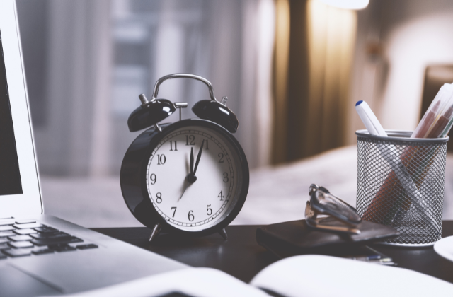 A black wind up clock on a dark desk next to to a laptop