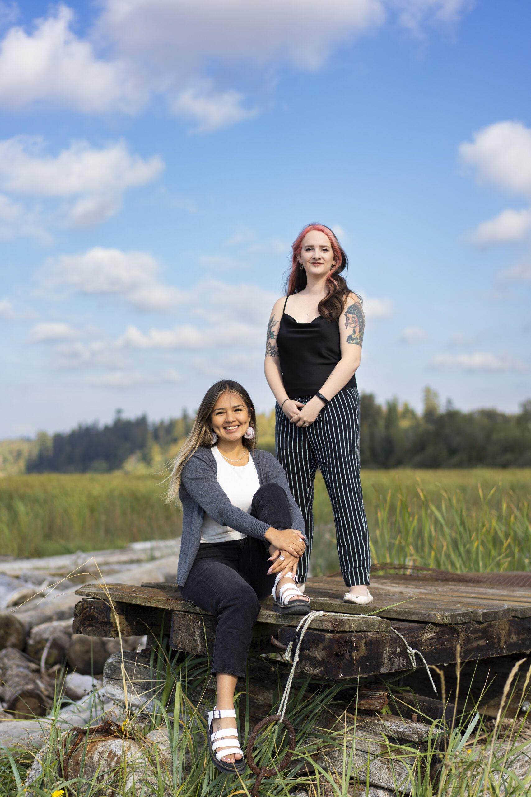 Qwu'yum'aat Elliott sits while Savannah Wells stands on a platform in a field posing, both portrayed as illustrious and vibrant.