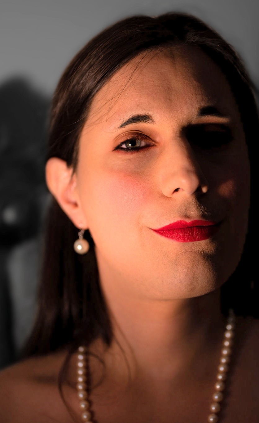 Woman wearing red lipstick and drop pearl earrings and bathed in soft lighting looks at the camera.