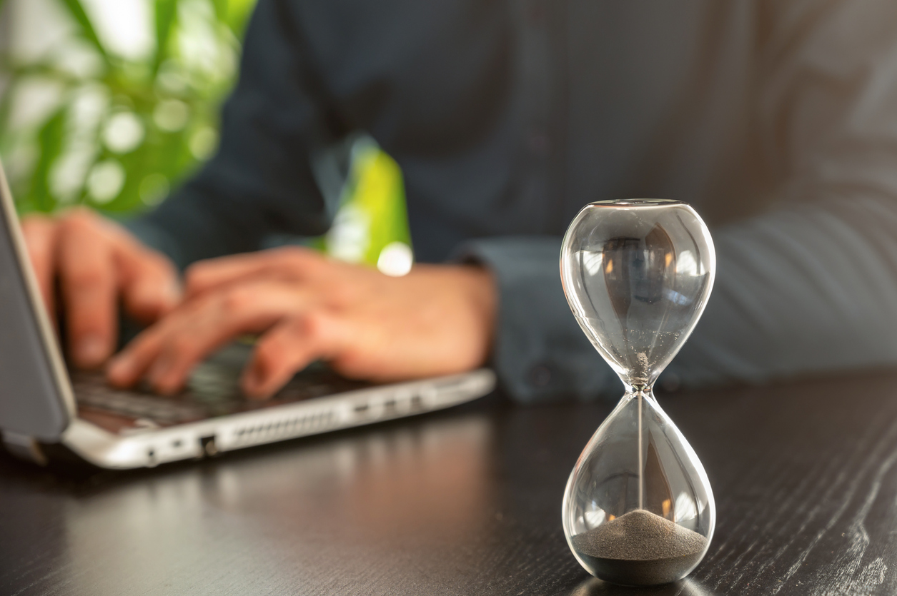 An hourglass with time running out in front of a person working on a laptop.