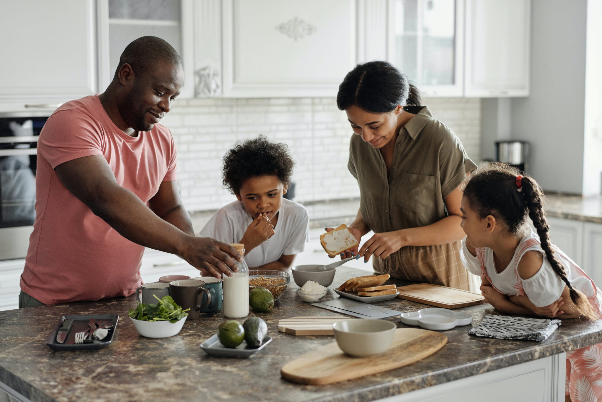 A family of four gather around their kitchen table passing food to each other. The father reaches for a bottle of milk, the mother spreads butter on her toast, and their two children are beside them smiling.