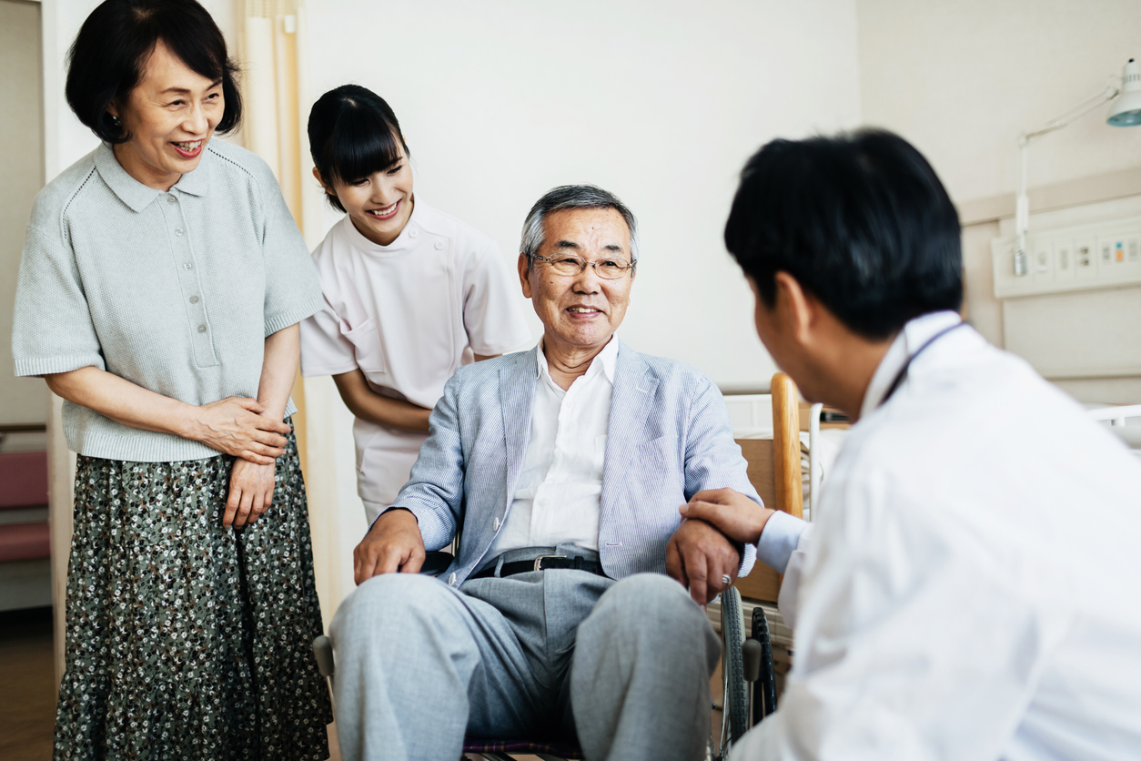 An elderly person is sitting in a wheelchair and talking with a doctor in front of him. His wife and a nurse stand behind him.
