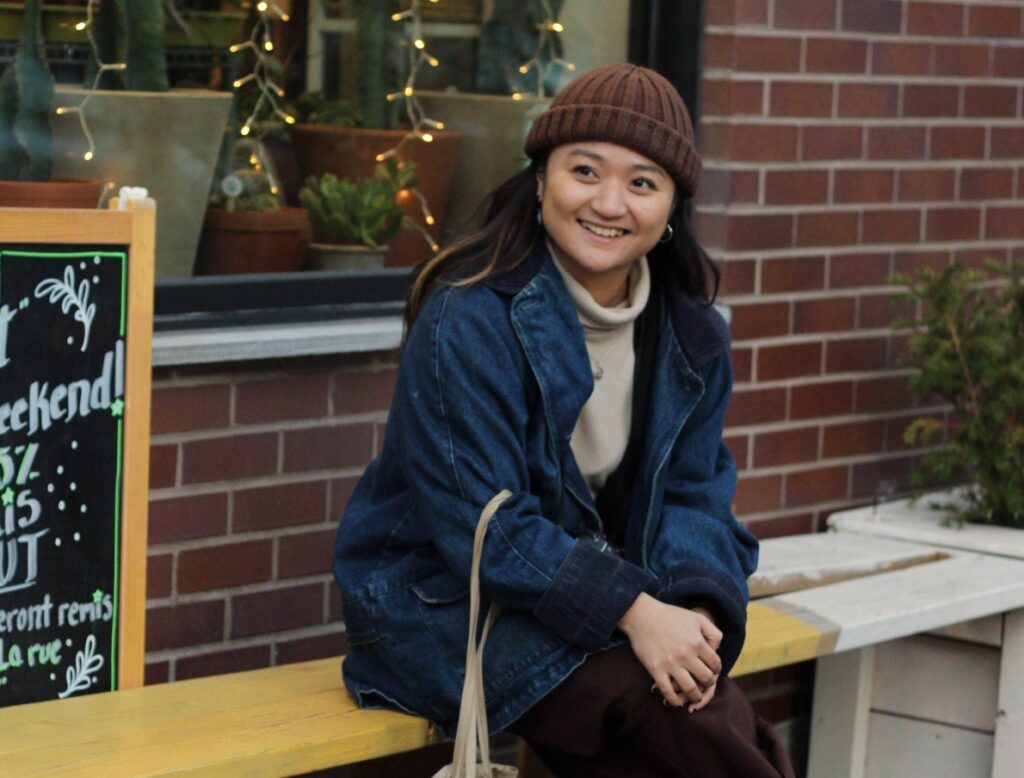 A person is pictured sitting on a bench, in front of a store front smiling.