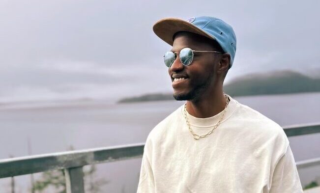 A person wearing sunglasses and a hat is pictured smiling in front of a scenic background.