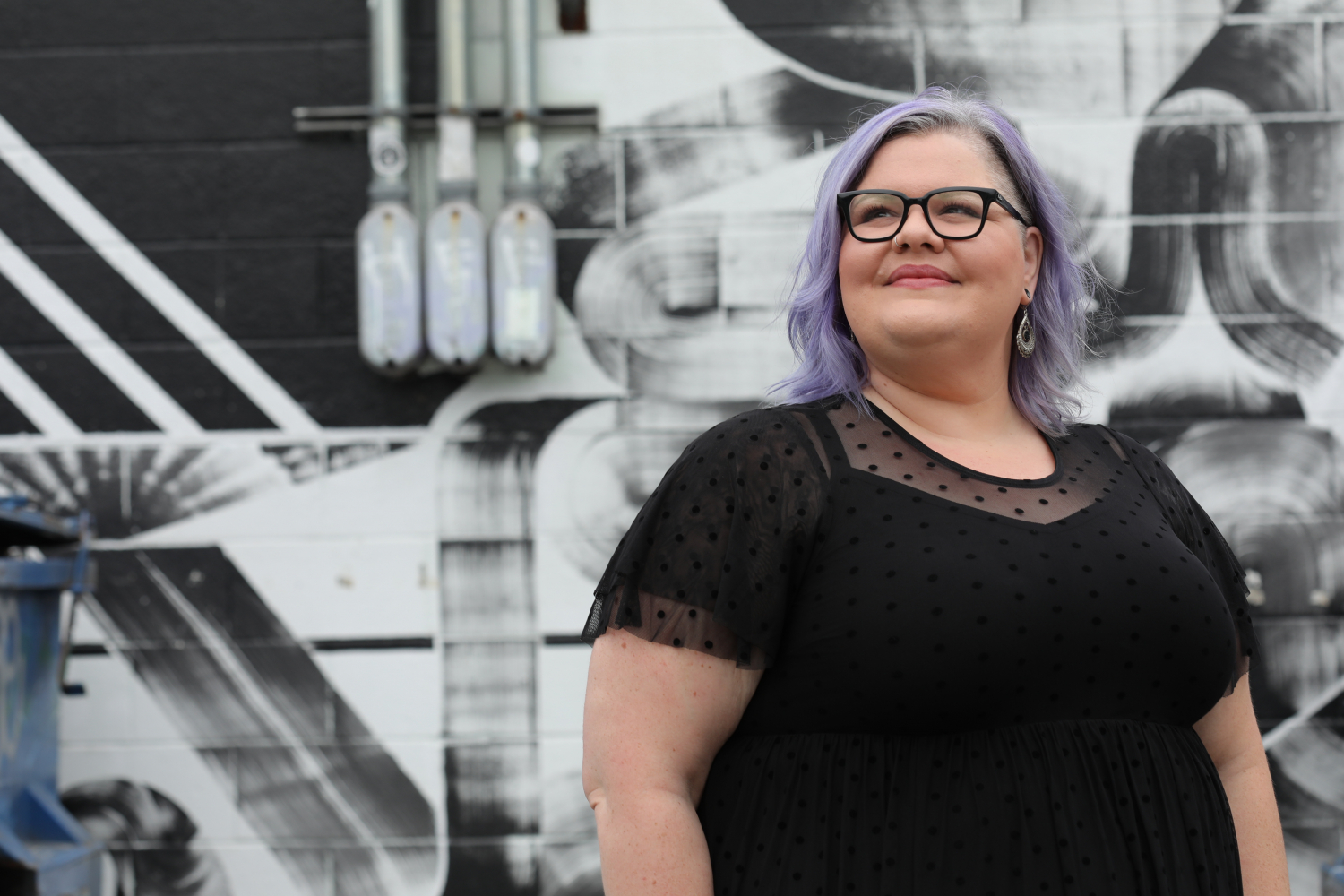 Smiling woman with lilac hair and dark glasses in a black polka dot top standing in front of a black and white mural