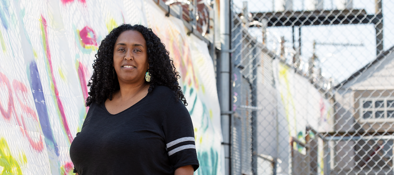 A smiling black woman in a black shirt standing near a colourful wall mural