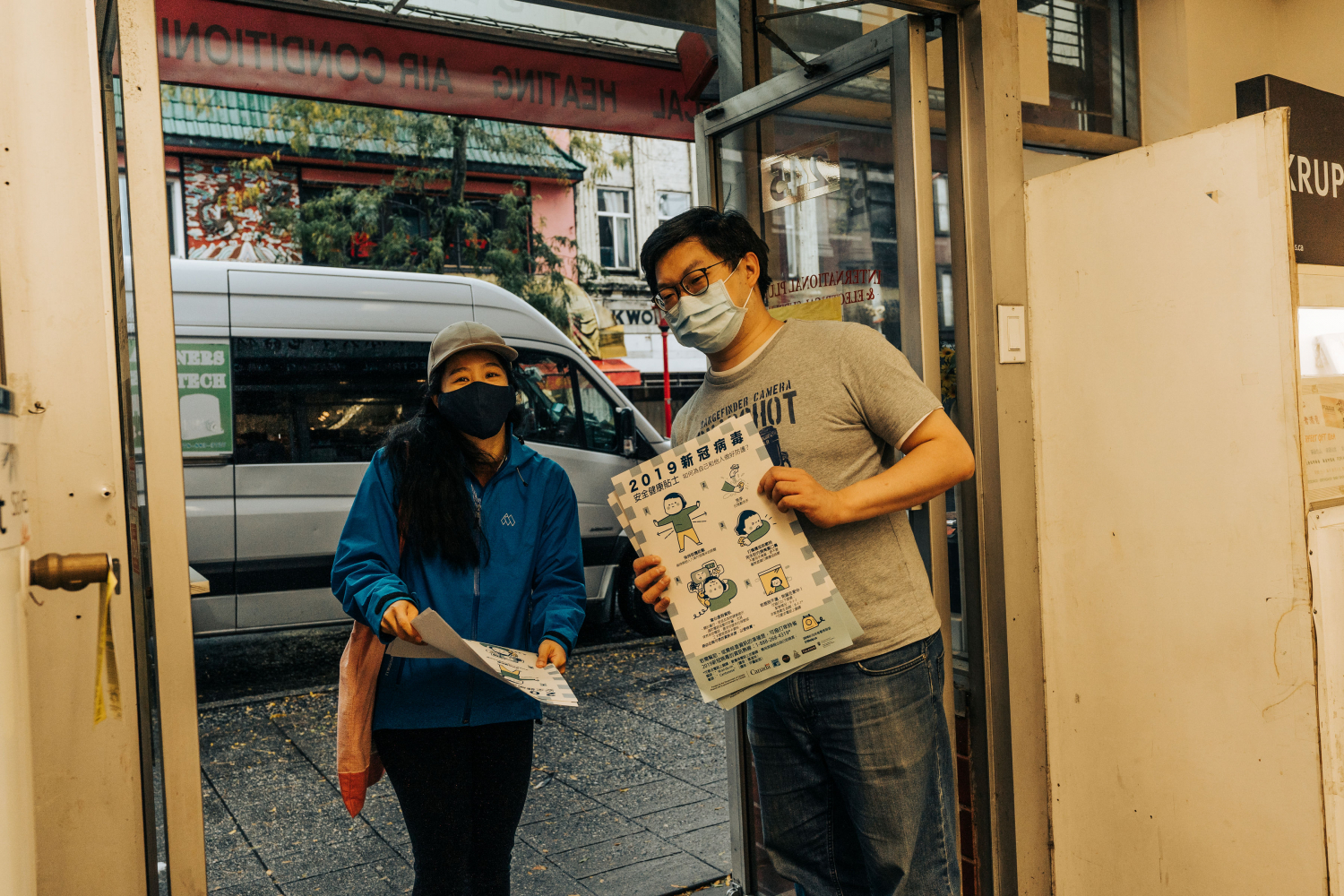 A smiling masked Asian man and woman, standing in a store doorway, holding posters