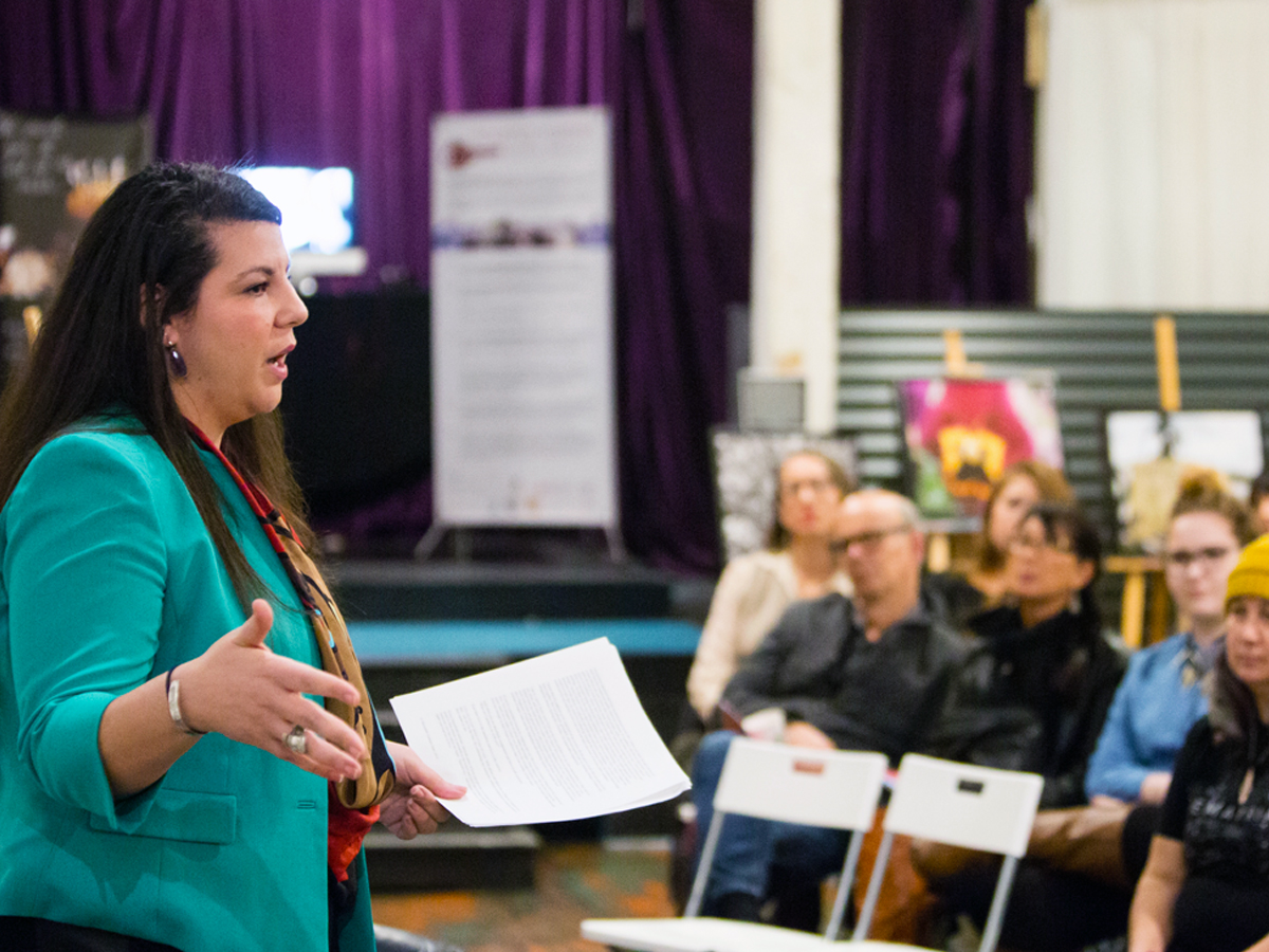 A woman with long brown hair and bright green blazer in profile, giving a lecture to a room of sitting people