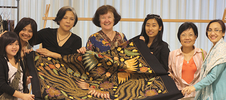 A diverse group of woman gathered around and displaying a dyed cloth artwork featuring birds and plants