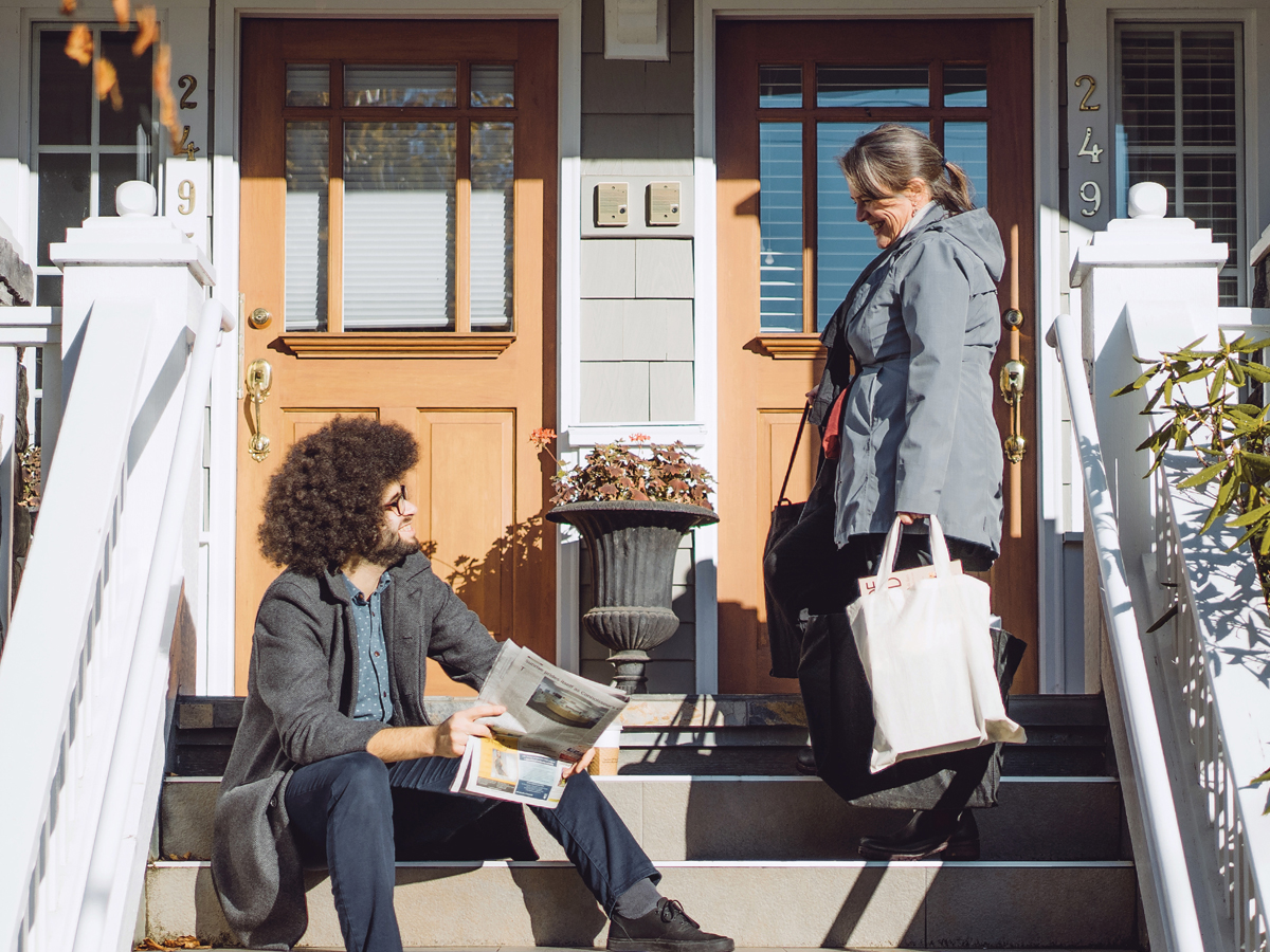 A man sitting on the steps of a townhouse holding a newspaper and smiling at a woman standing on the steps holding grocery bags