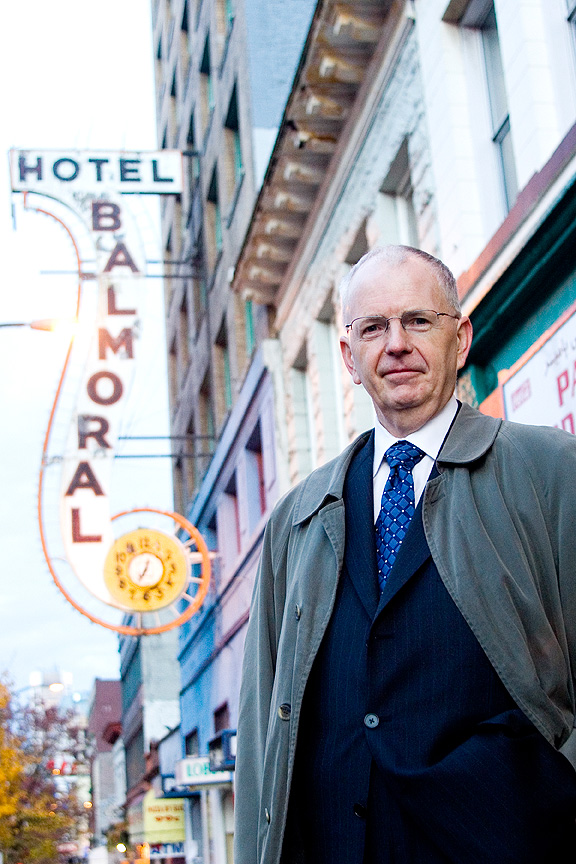 An older man in glasses, suit and jacket, standing, with a sign in the background reading "Hotel Balmoral"