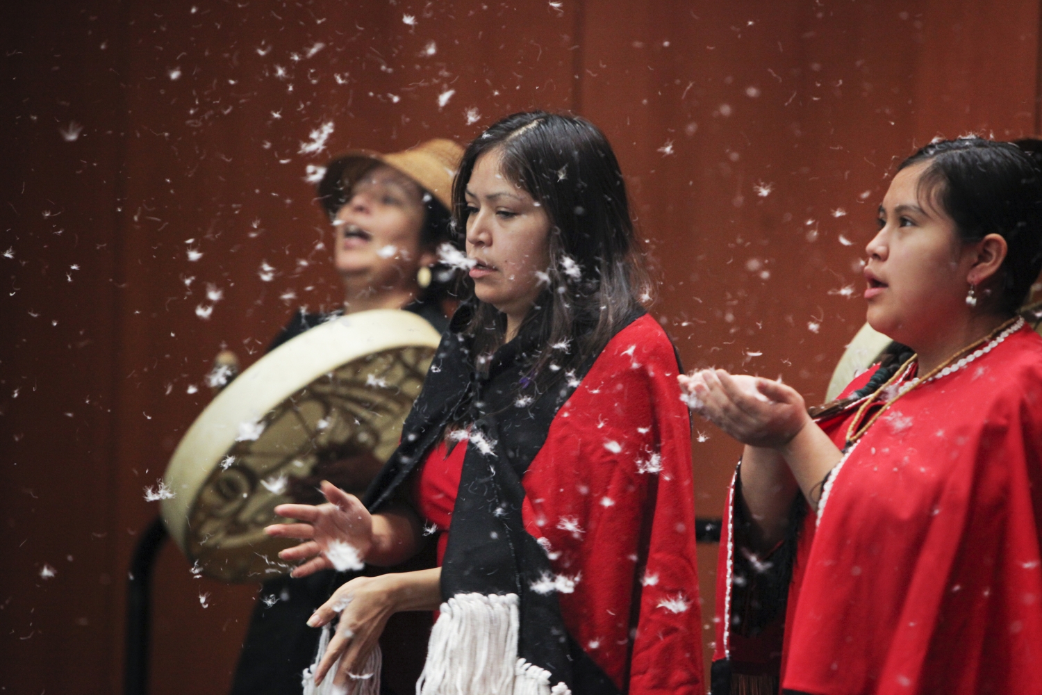 Two Indigenous women performers in black and red cloaks catching falling feathers in cupped hands as another performer in a woven hat plays a hand drum in the background