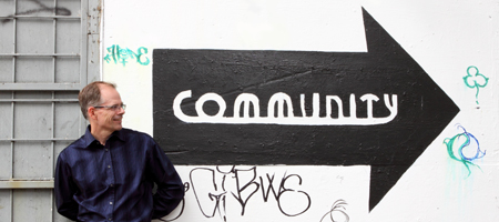 IMG_1238_WEB_0 Man leaning on a graffitied wall with a black arrow pointing right painted on it and the word "Community" in white inside the arrow
